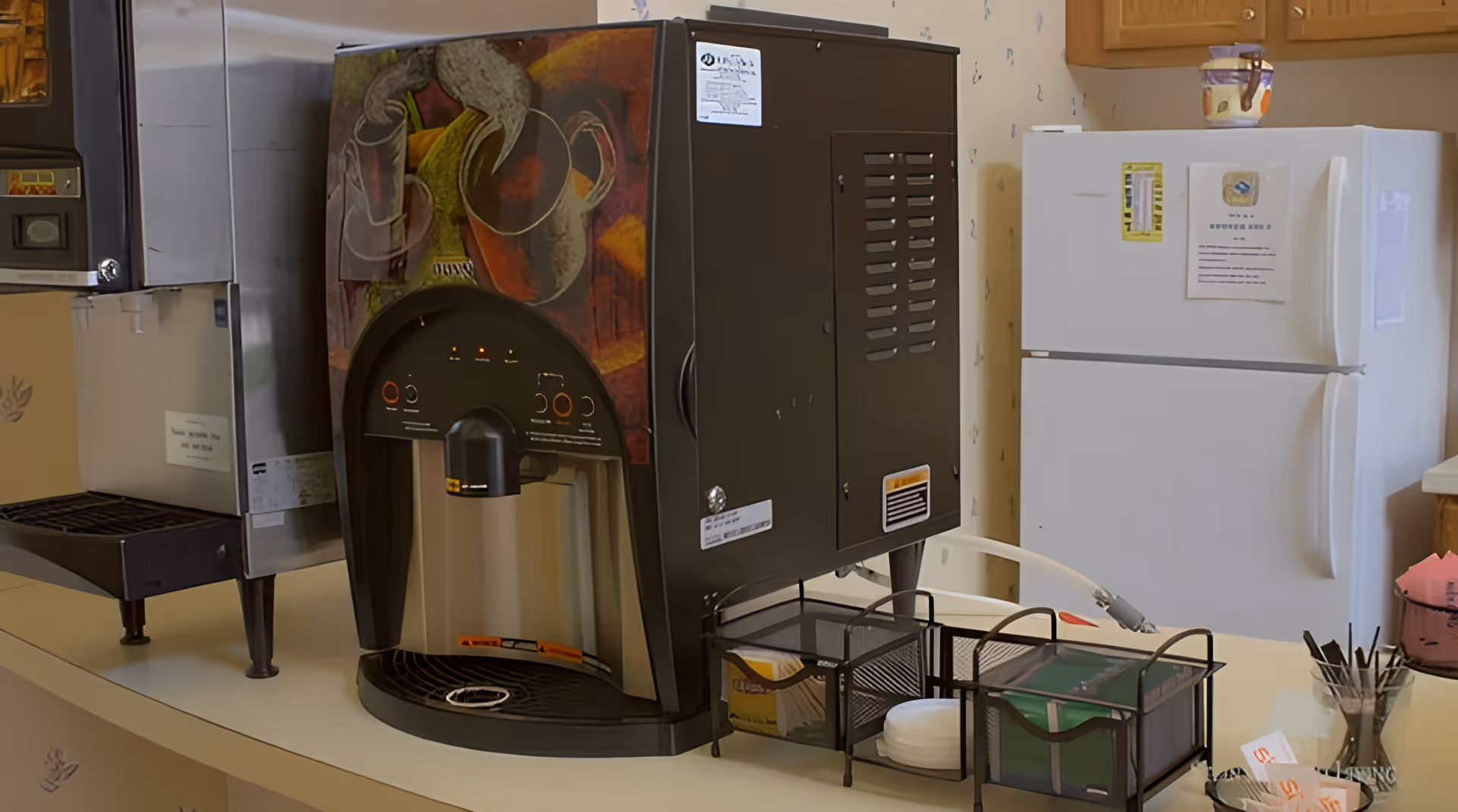 A coffee and beverage station in a kitchen area featuring a large coffee machine with artistic designs on the side, a white refrigerator with papers and notes attached, and various containers holding stirrers, napkins, and sugar packets on the countertop.