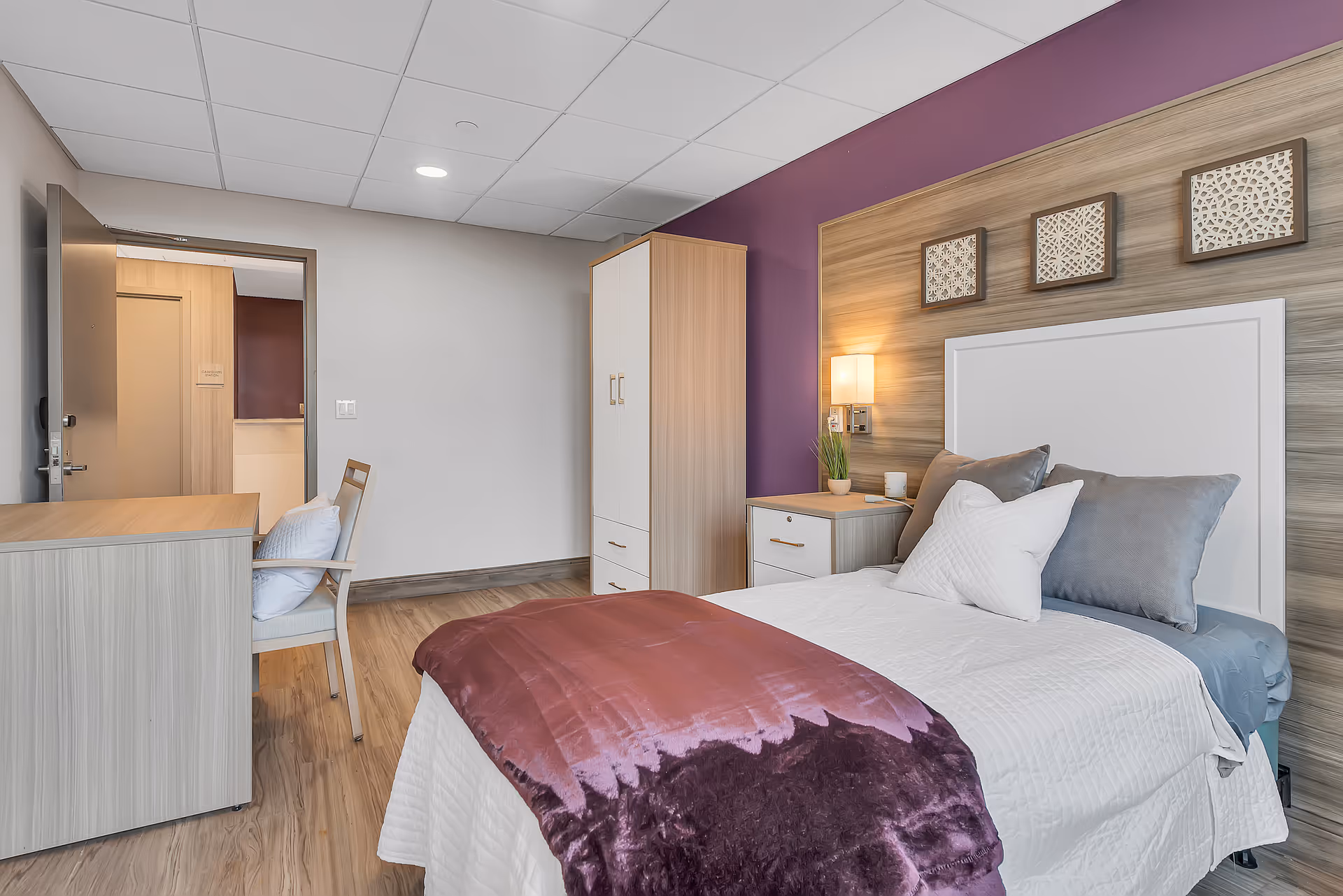A cozy bedroom in The Veranda Assisted Living featuring a neatly made bed with white and gray pillows and a purple throw blanket. The room has a wooden floor, a desk with a chair, a wardrobe, and a nightstand with a lamp and a small plant. The wall behind the bed is decorated with three framed art pieces and painted purple.