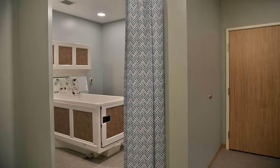A small room with a medical or therapeutic bathtub partially hidden behind a patterned privacy curtain. The walls are painted light gray, and there is a wooden door on the right side of the image. The room is well-lit with a ceiling light.