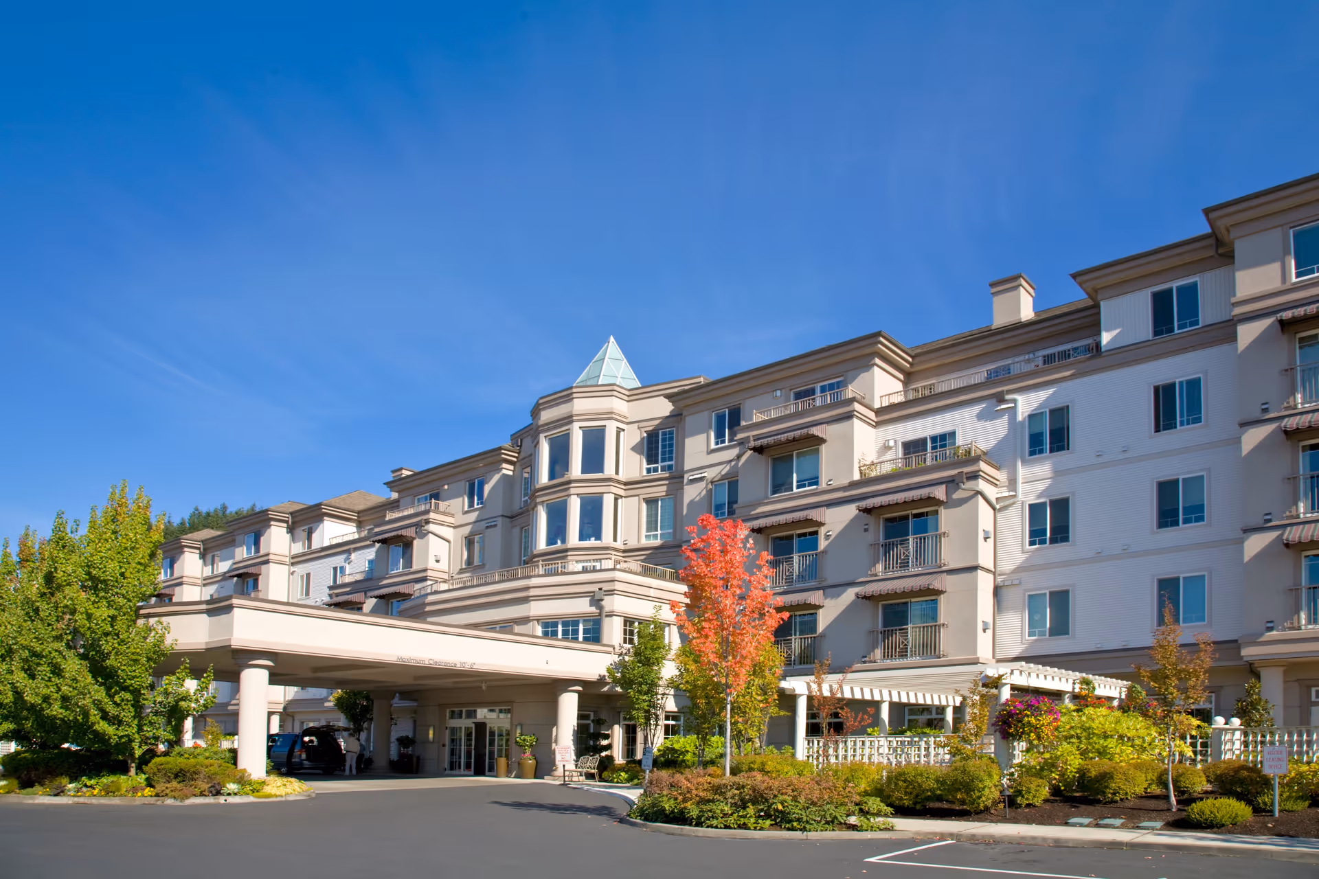 Exterior view of a multi-story senior living facility building under a clear blue sky, with a covered entrance, landscaped greenery, and a tree with red autumn leaves in front.