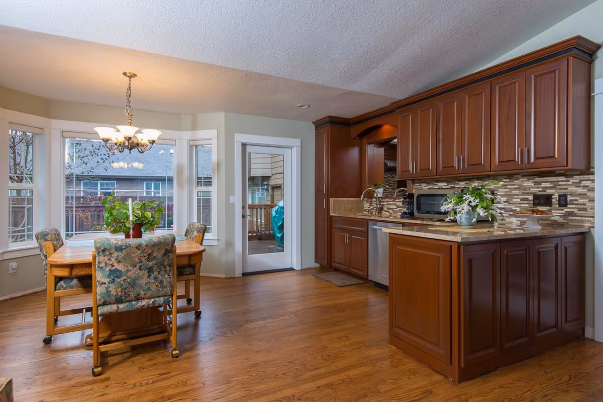 A bright kitchen and dining area with wooden floors and cabinetry. The kitchen features dark wood cabinets, a countertop with a vase of flowers, a microwave, and a dishwasher. The dining area has a wooden table with four floral upholstered chairs, a chandelier overhead, and large windows providing natural light and a view of the outdoor deck and neighboring houses.