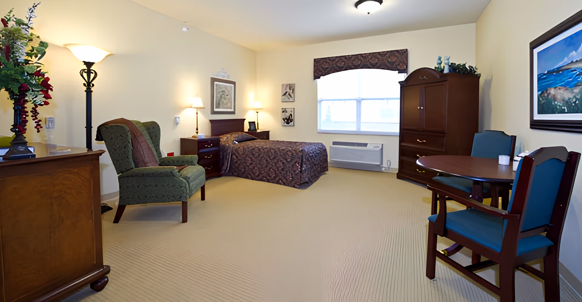 Spacious furnished senior living studio with a bed, armchair, armoire, dresser, and a small dining table under soft lighting.