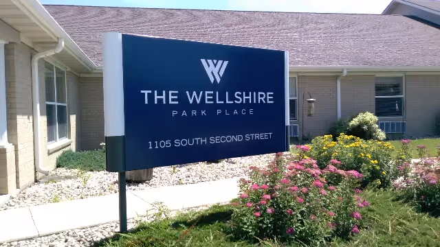 Outdoor view of The Wellshire Park Place facility sign with the address 1105 South Second Street, surrounded by flowering plants and bushes in front of a single-story building.
