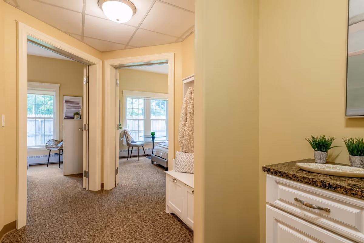 View down a hallway in a senior living facility with two open doorways leading to bedrooms. Each bedroom has large windows letting in natural light, with chairs and beds visible. The hallway has beige walls and carpeted floors, with a white cabinet topped with a granite countertop and small potted plants on the right side.