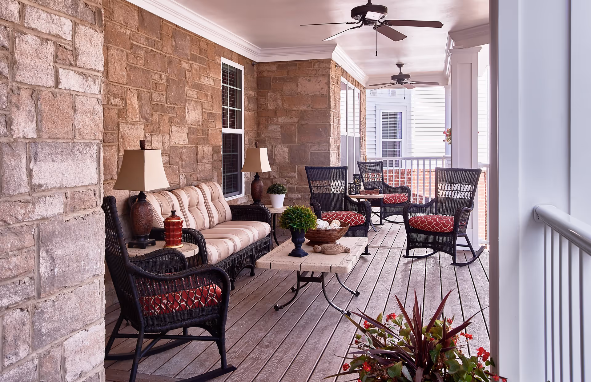 A covered outdoor porch area with stone walls and wooden flooring, furnished with a cushioned sofa, four wicker rocking chairs with red patterned cushions, two table lamps, a coffee table with decorative items, ceiling fans, and potted plants.