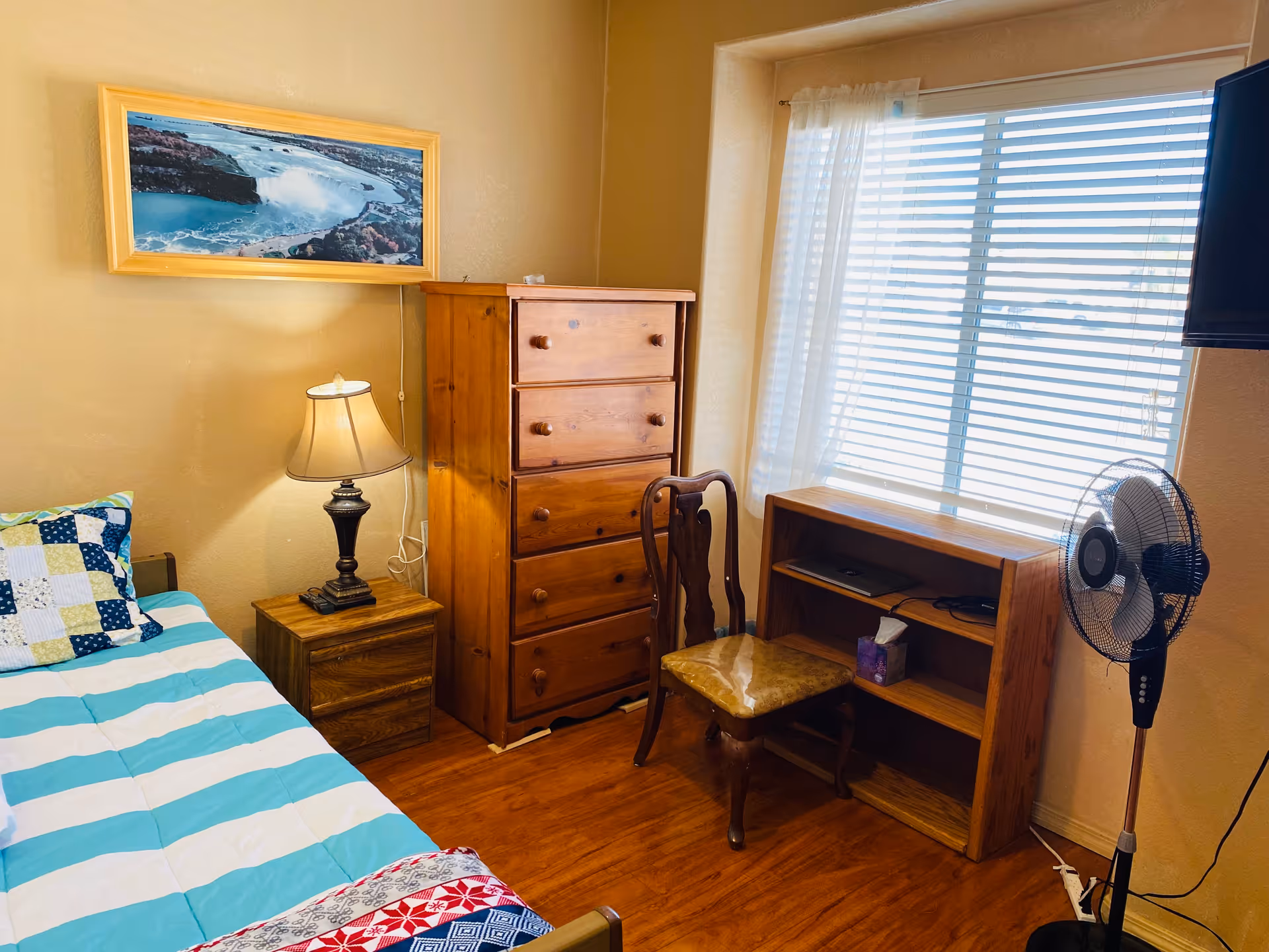 A cozy bedroom with a single bed covered in a striped blue and white blanket and a patchwork pillow. Next to the bed is a wooden nightstand with a lamp. There is a tall wooden dresser against the wall and a wooden chair with a cushioned seat in front of a small wooden bookshelf. A standing fan is positioned near a window with white blinds and sheer curtains. A framed picture of a waterfall hangs on the wall above the nightstand.