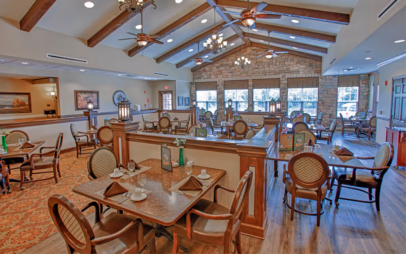 Spacious dining room with multiple set tables and wooden chairs under a vaulted beamed ceiling and large windows.