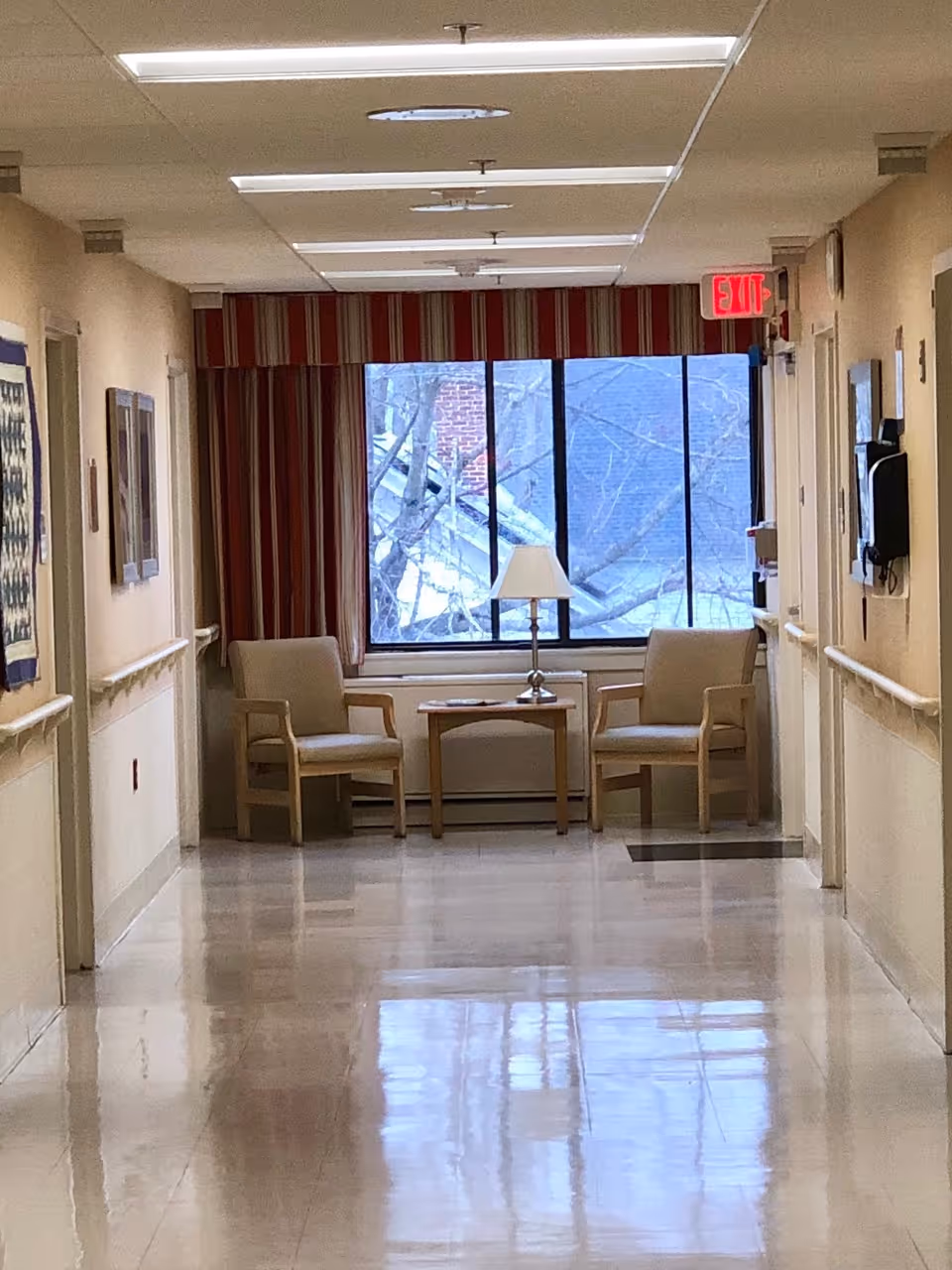 Polished hallway in a care facility leading to a seating area with two chairs, a small table and lamp in front of a large window.