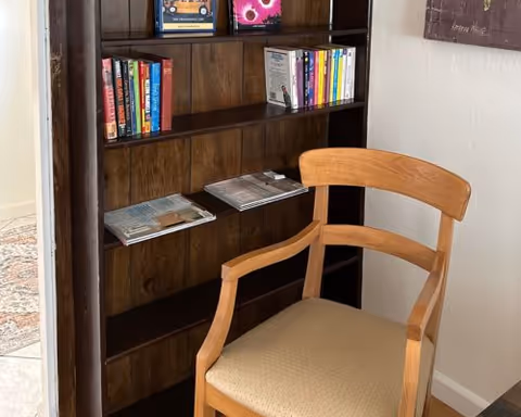 Wooden chair positioned in front of a dark wood bookshelf holding books and magazines in a home's interior.