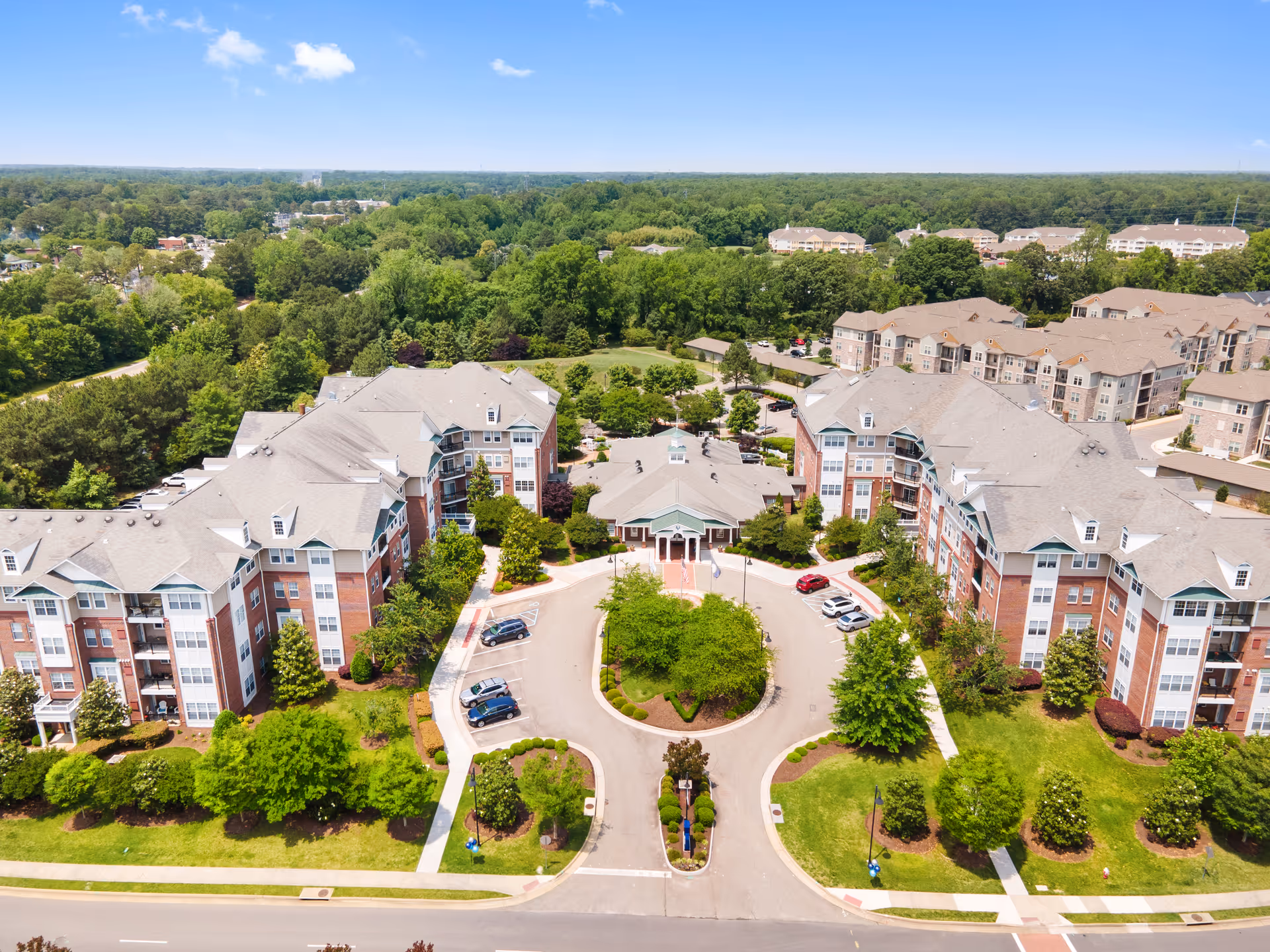Aerial view of Verena at the Reserve senior living facility showing multiple large residential buildings arranged around a circular driveway with landscaped greenery and trees. The buildings have multiple stories with gray roofs and red brick facades. Surrounding the facility are dense green trees and a clear blue sky.