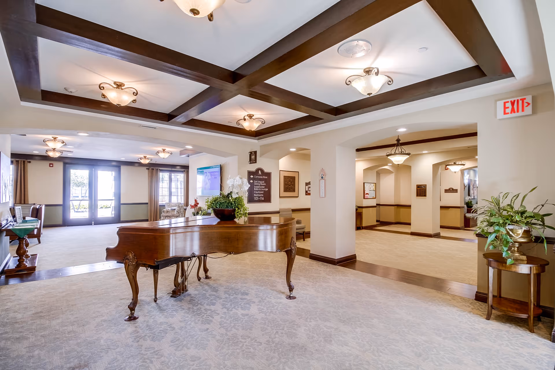 Spacious and well-lit interior common area with a grand piano in the center, decorative ceiling beams with multiple light fixtures, beige carpet, and walls painted in neutral tones. There are plants on small tables, a signboard on the wall, and large windows and doors letting in natural light.