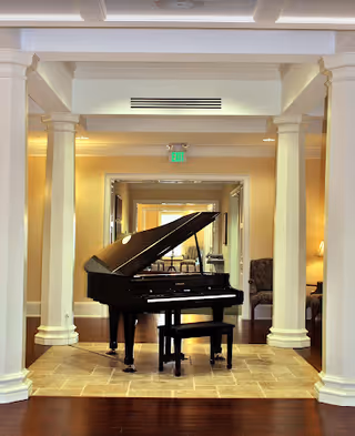 A grand piano on a tiled platform in a well-lit assisted living facility common area framed by columns and seating.