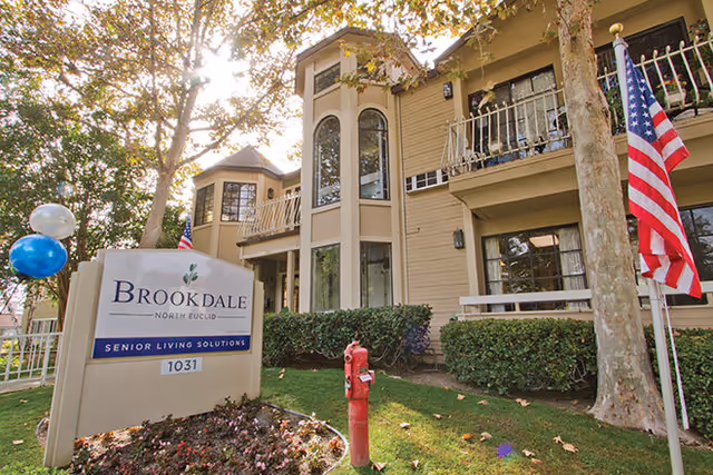 Exterior view of Brookdale North Euclid senior living facility showing a multi-story beige building with balconies, surrounded by trees and greenery. In the foreground, there is a sign with the facility name and address, along with a red fire hydrant and an American flag.
