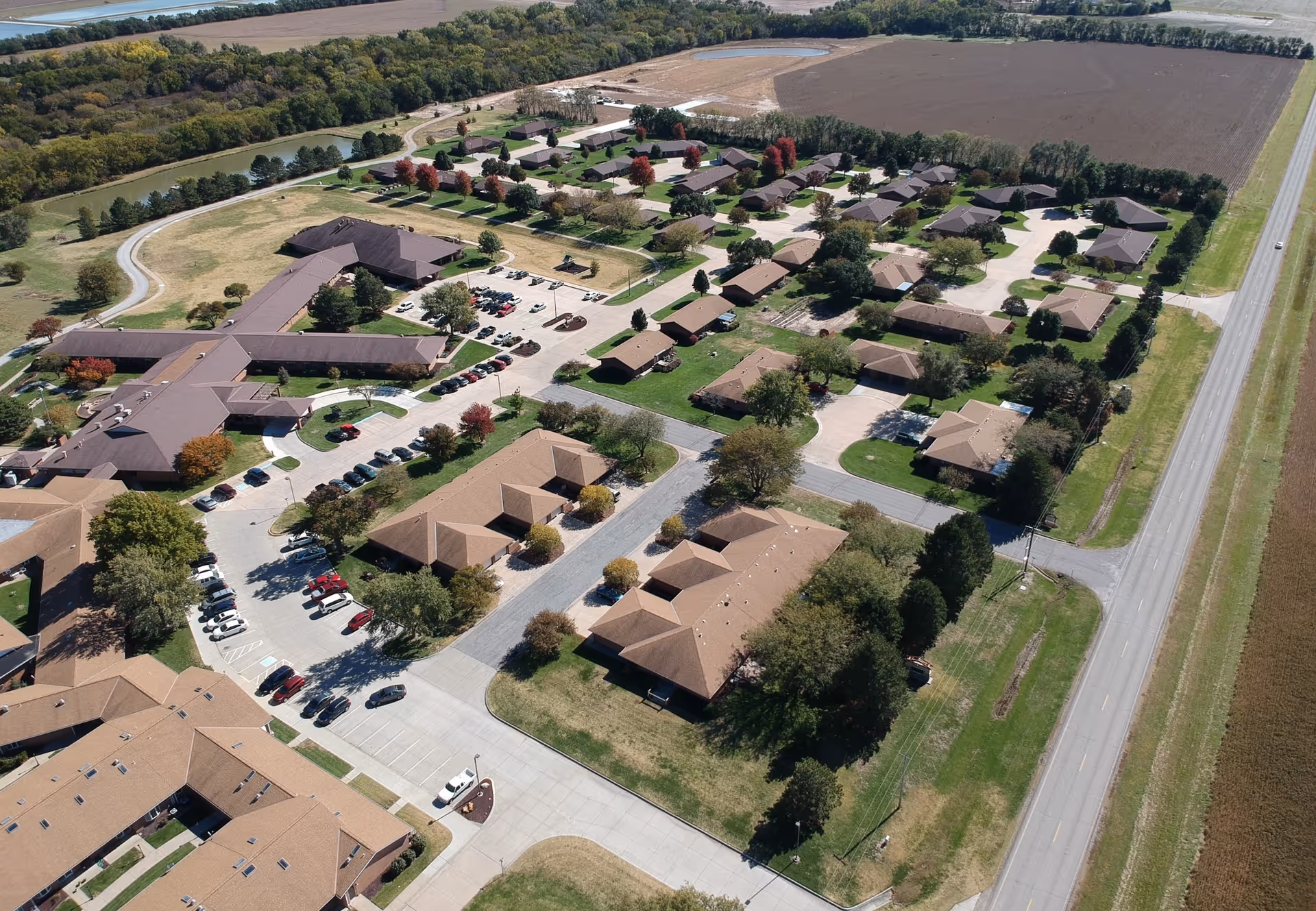Aerial view of Pine Village senior living facility showing multiple single-story buildings with brown roofs, parking lots with cars, green lawns, trees, and surrounding farmland.