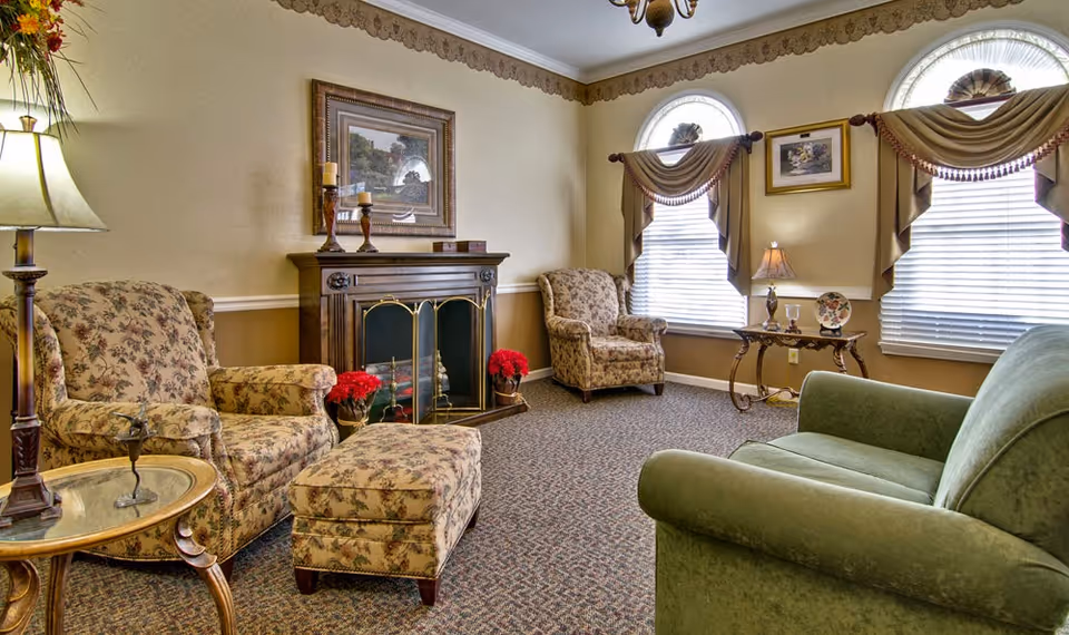 A cozy living room with floral upholstered armchairs and matching ottoman, a green sofa, a wooden fireplace with decorative candles, two arched windows with draped curtains, a side table with a lamp and decorative items, and a carpeted floor.
