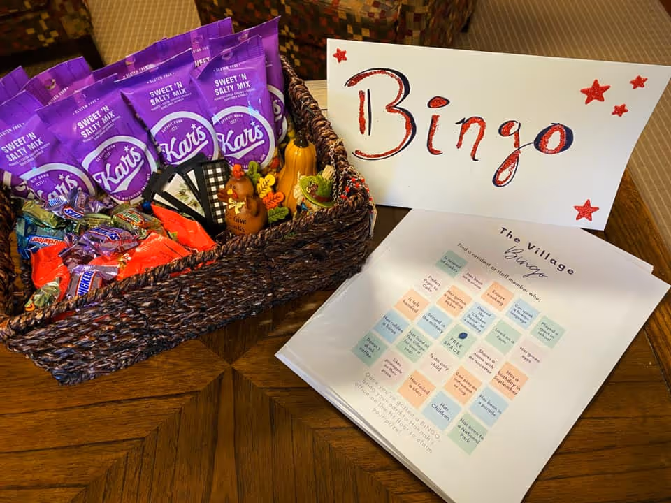 A woven basket filled with snack bags including Kar's Sweet 'N Salty Mix and various candy bars, placed on a wooden table next to a handmade sign that says 'Bingo' decorated with red stars, and a stack of bingo cards titled 'The Village Bingo'.