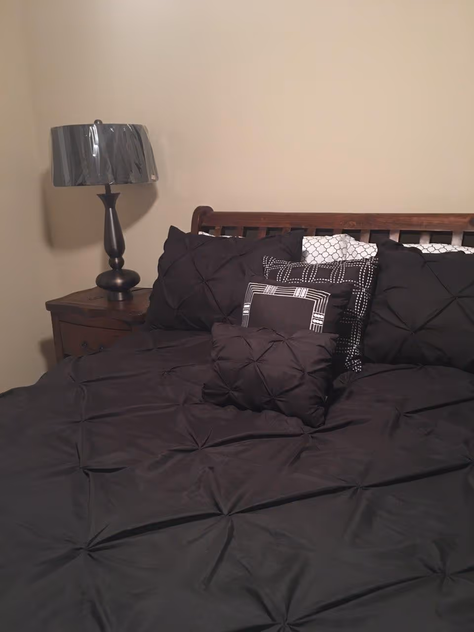 A neatly made bed with a dark comforter and multiple decorative pillows in black and white patterns. Next to the bed is a wooden nightstand with a dark-colored lamp with a shiny lampshade. The wall behind the bed is plain and beige.