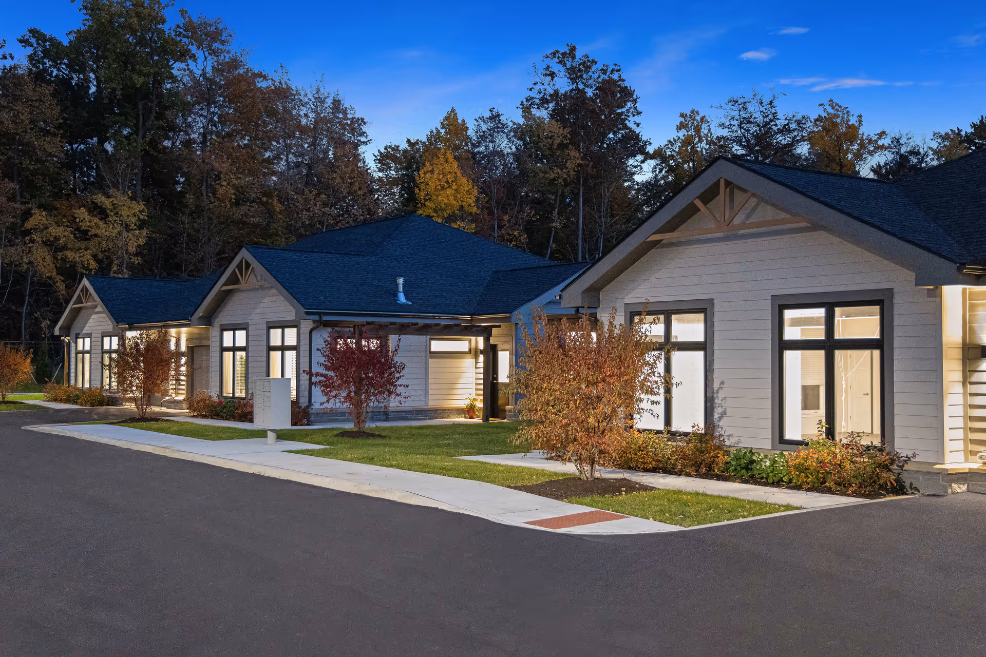 Exterior view of a single-story building with multiple sections, large windows, and a dark roof, surrounded by small trees and shrubs, with a paved driveway and sidewalk in front during dusk.