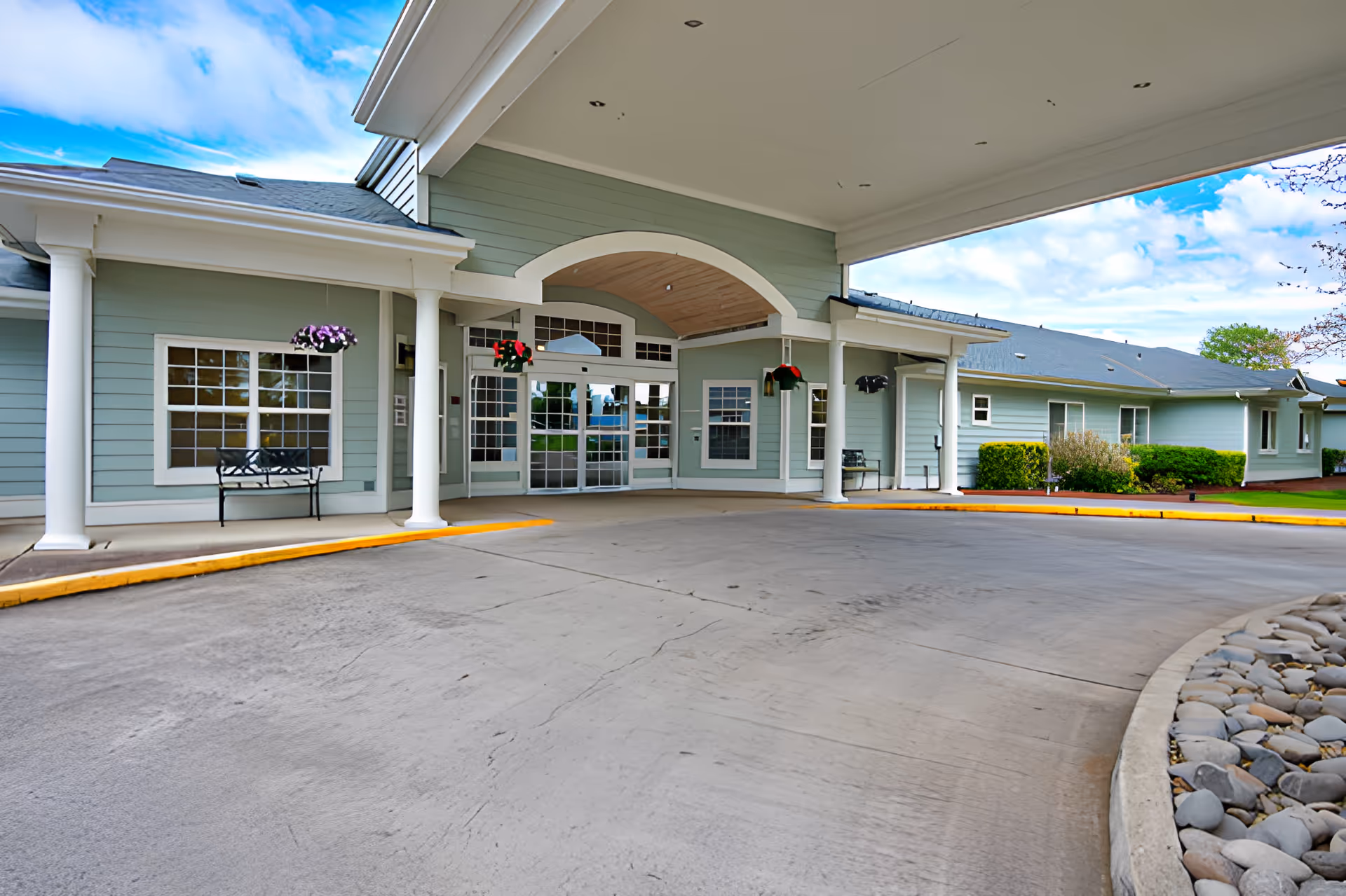 Covered entrance of a building with light green siding, white columns, and large glass doors. There are hanging flower baskets and benches near the entrance. The driveway curves around a landscaped area with rocks and bushes.