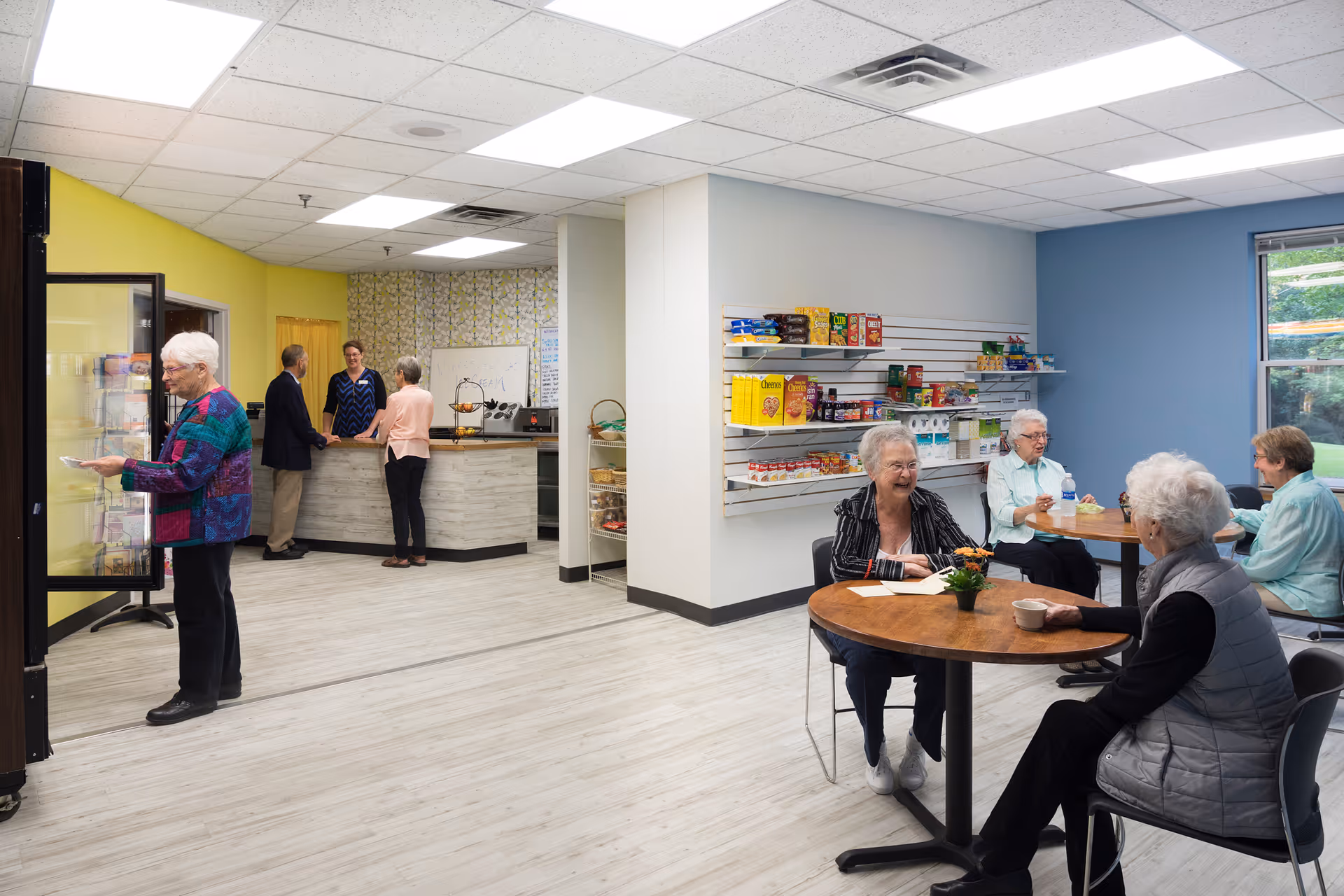 A bright and clean common area in a senior living facility where elderly residents are socializing. On the right, four seniors are seated around two round wooden tables, chatting and enjoying drinks. Behind them, shelves stocked with various snacks and food items are mounted on a light blue wall. On the left side, a woman is selecting items from a refrigerated display case, while two other seniors are conversing with a staff member behind a counter with a yellow wall background. The floor is light-colored wood, and the ceiling has recessed lighting panels.
