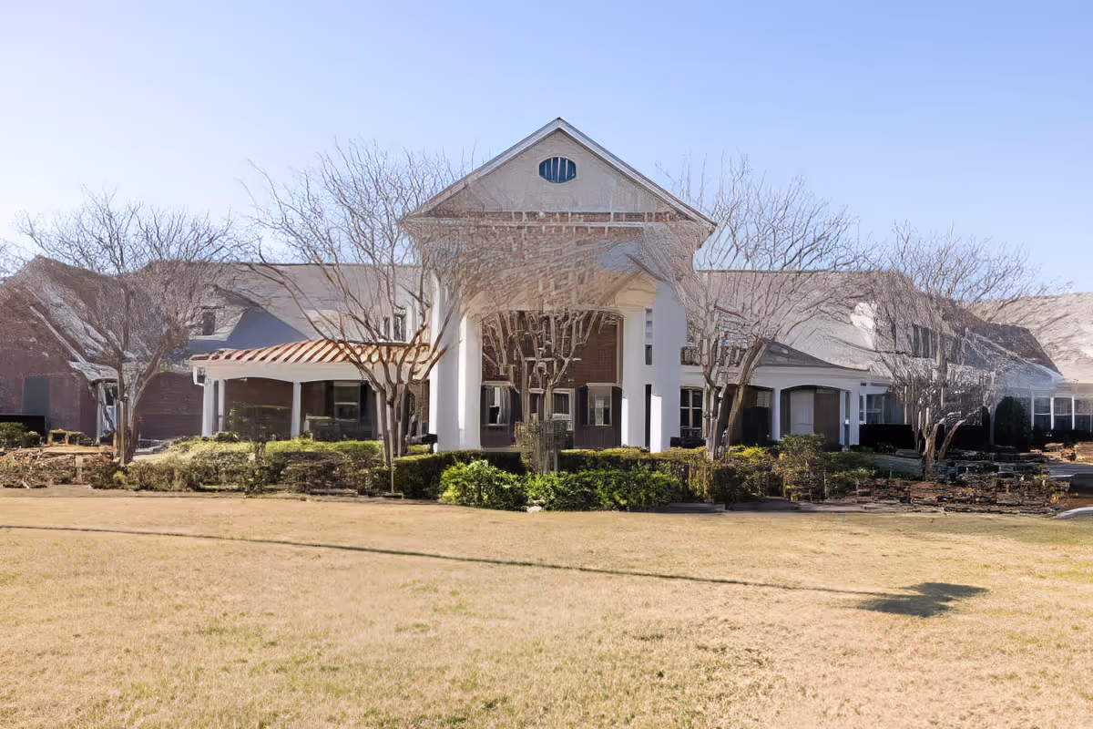 Front exterior of a single-story senior living facility with a columned entrance, manicured shrubs, and a grassy lawn.