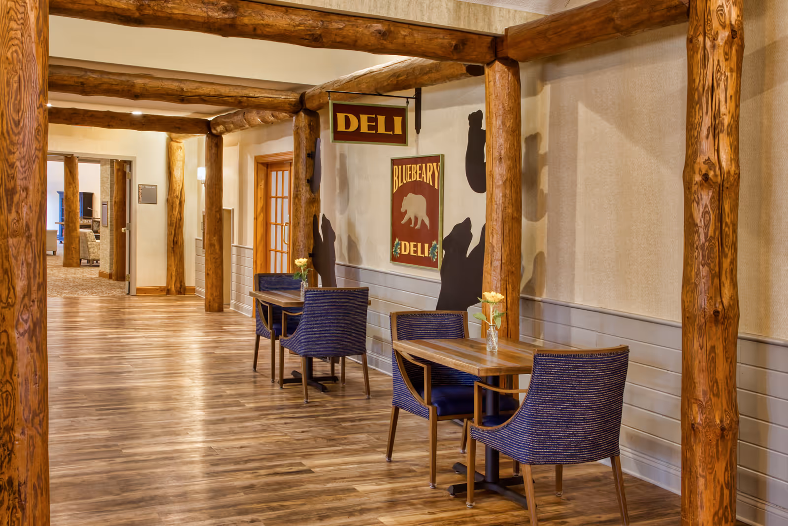 Interior hallway of a senior living facility with wooden log beams and columns. Two small tables with blue chairs and vases with yellow flowers are placed along the wall. Signs reading 'DELI' and 'BLUEBERRY DELI' with a bear graphic hang on the wall. The floor is wooden and the hallway leads to another room with seating visible in the background.