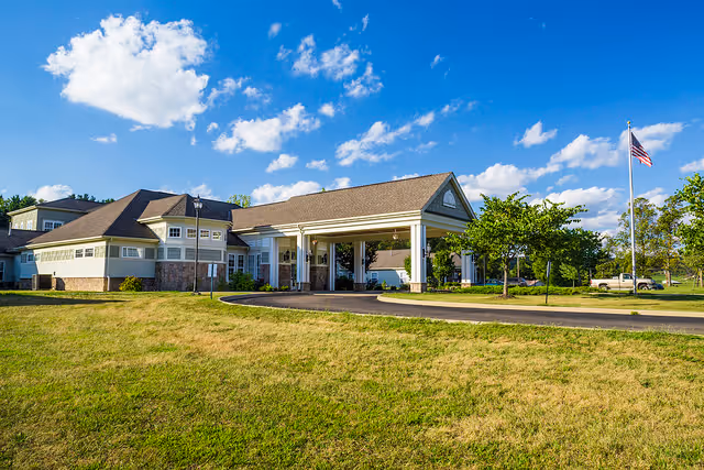 Exterior view of a single-story senior living facility building with a covered entrance, surrounded by a grassy lawn and trees under a blue sky with scattered clouds. An American flag is visible on a flagpole to the right.
