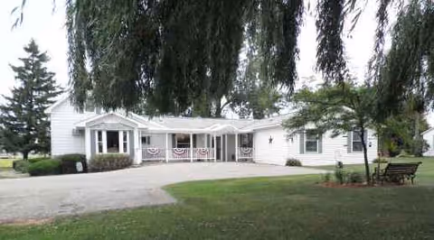 Single-story white retirement facility with a front entrance, porch railing, and neatly kept lawn.