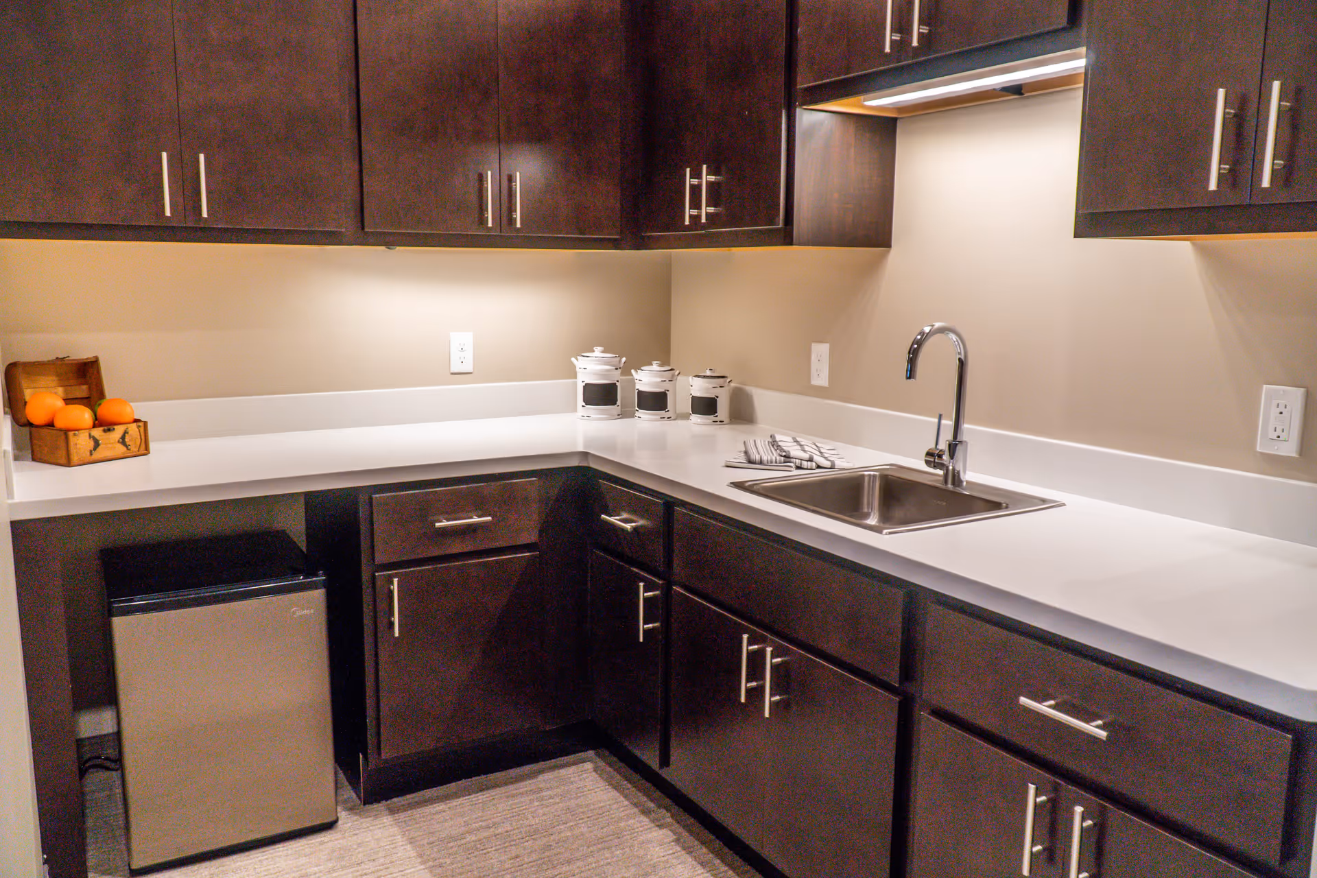 A modern kitchen area with dark wood cabinets, a white countertop, a stainless steel sink with a faucet, a small stainless steel mini fridge, three white canisters, a striped dish towel, and a small wooden crate holding oranges.