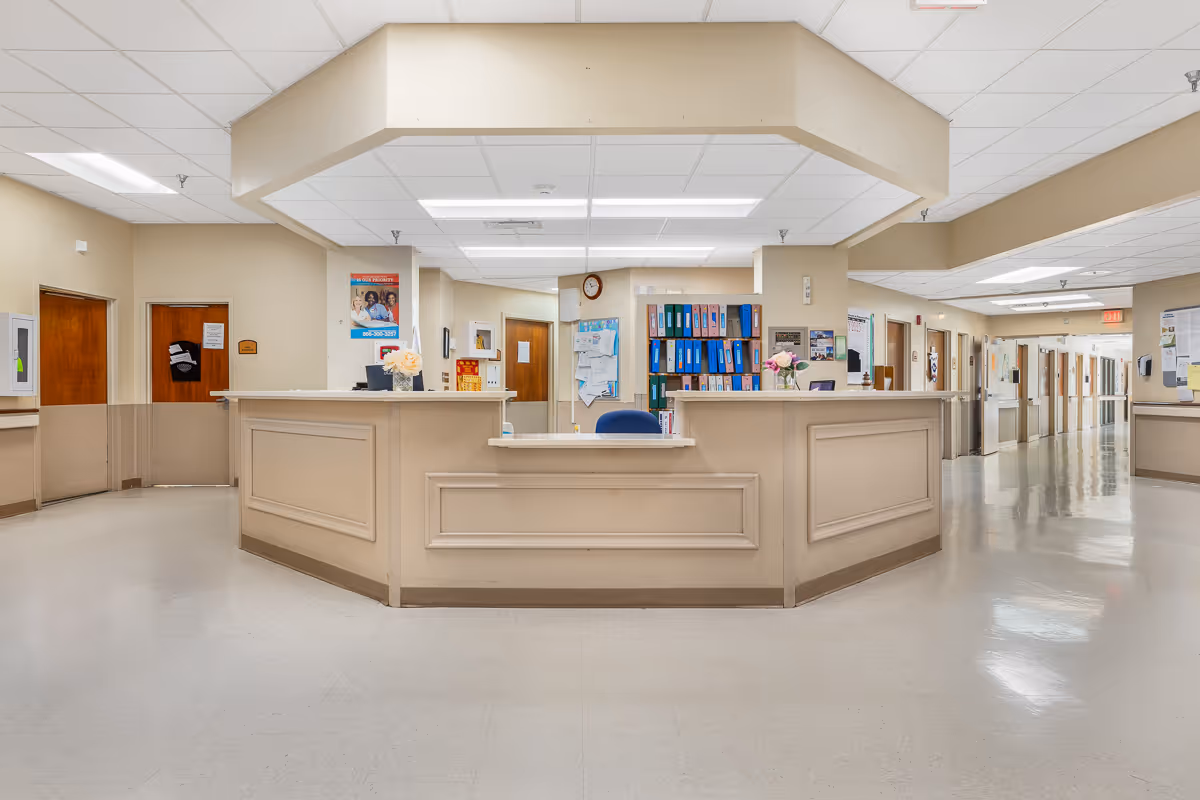 A central nurses' station/reception desk in a bright interior hallway of a senior healthcare facility, with filing shelves, doors, and bulletin boards along the corridor.