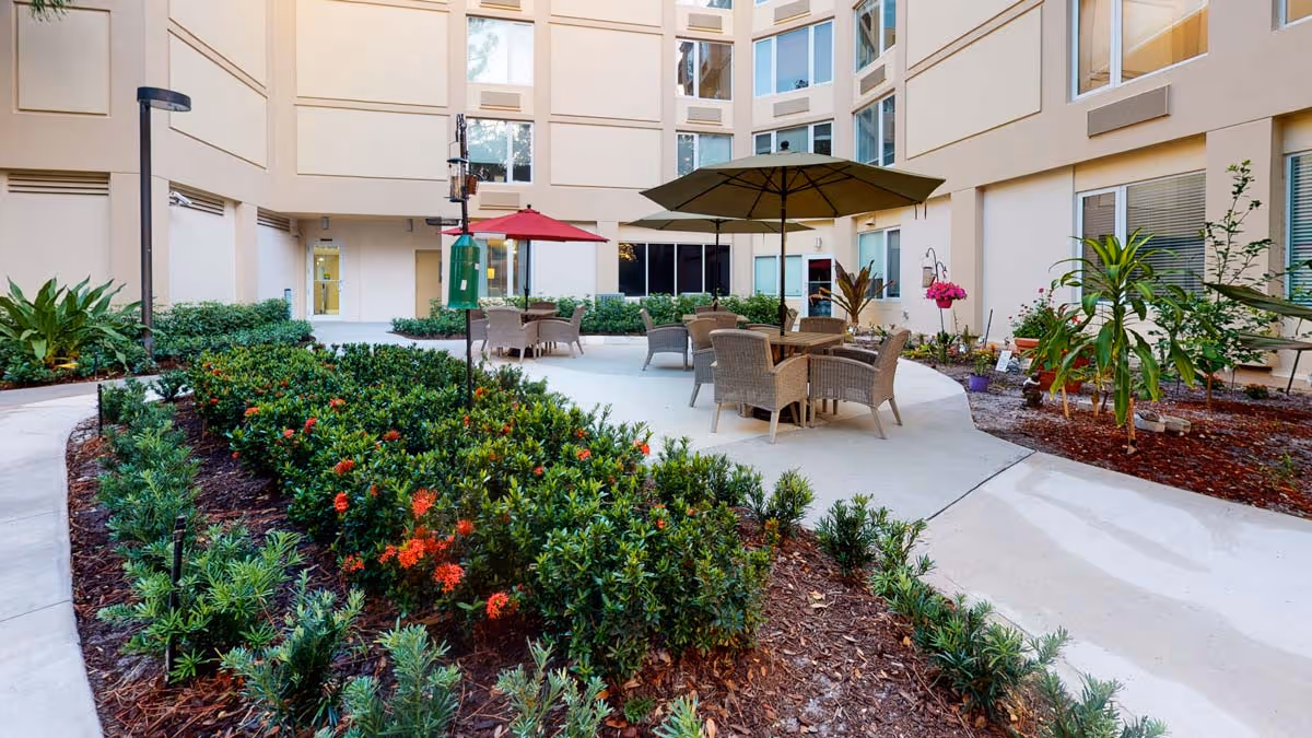 Outdoor courtyard area at The Atrium at Boca Raton featuring a paved seating area with tables, chairs, and umbrellas surrounded by landscaped garden beds with green shrubs and flowering plants, enclosed by the building's beige exterior walls and windows.