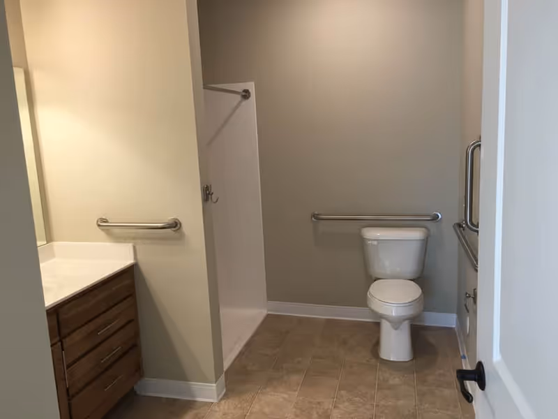 A clean and accessible bathroom featuring a white toilet with grab bars on both sides, a walk-in shower with a grab bar and a hook, a vanity with a white countertop and wooden drawers, and beige tiled flooring.