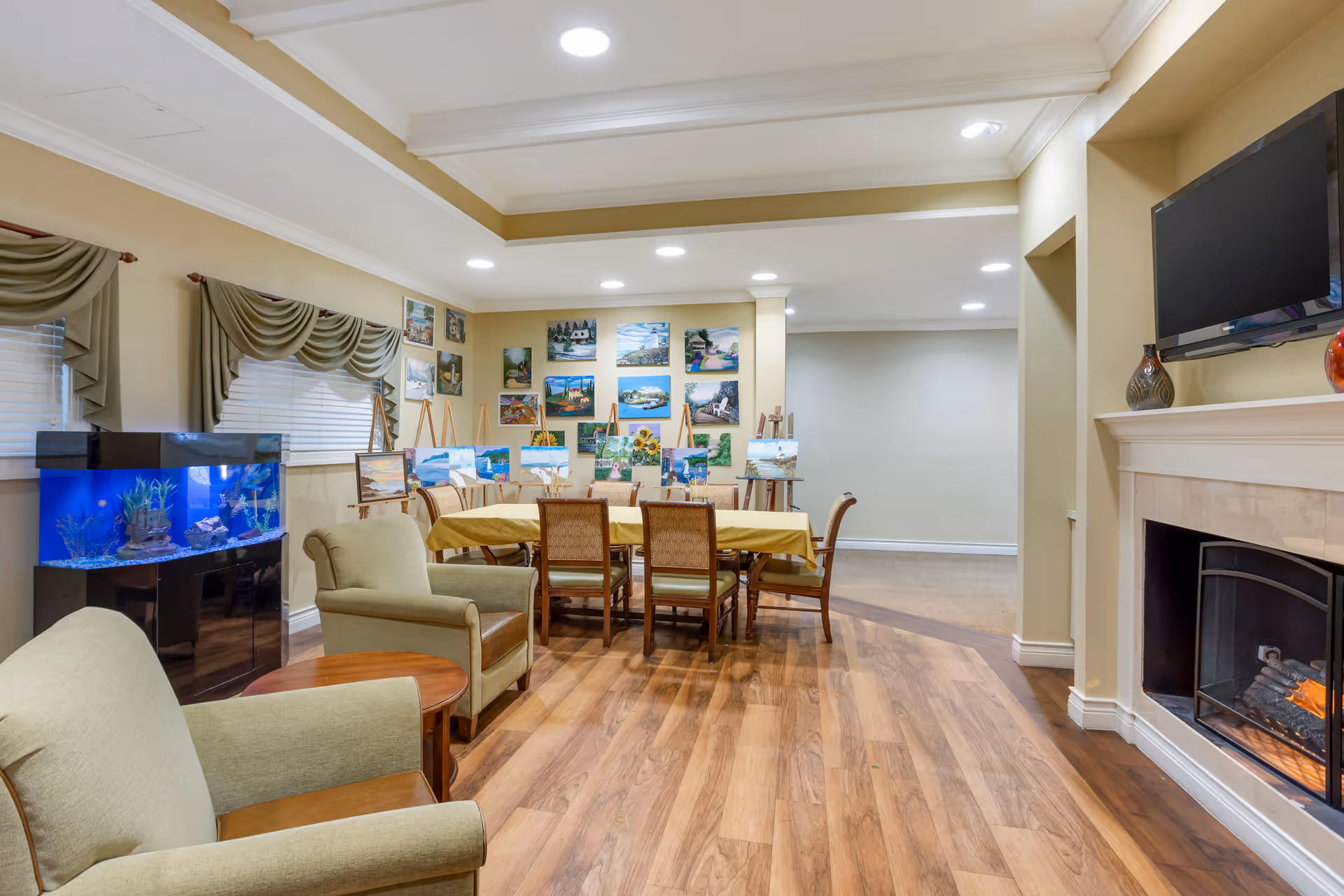 A bright common room with a dining table and chairs, armchairs, an aquarium, fireplace, and artwork displayed on easels and walls.