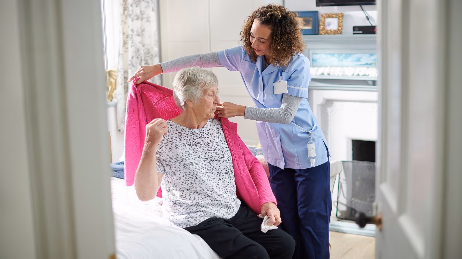 A caregiver helps an elderly woman put on a bright pink cardigan while sitting on a bed in a cozy, well-lit bedroom with a fireplace and decorative items in the background.