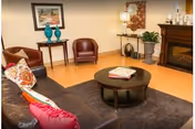 A cozy living room with a dark brown leather sofa adorned with colorful pillows, a round dark wood coffee table with books on it, two brown leather chairs, a small table with decorative blue vases, a floor lamp, a fireplace, and framed artwork on the walls.