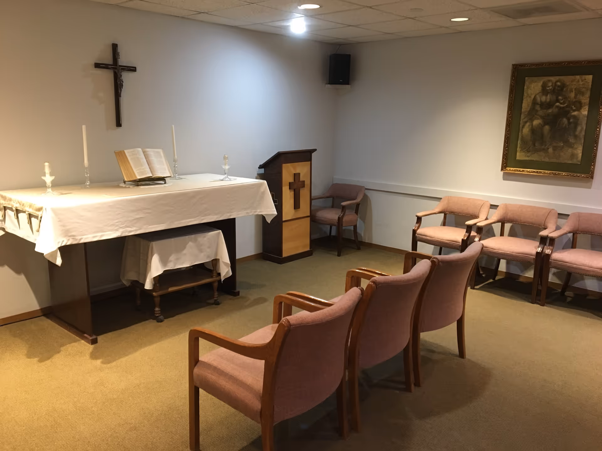 Small chapel-like interior with an altar covered in a white cloth, a crucifix on the wall, a lectern, and rows of pink upholstered chairs.