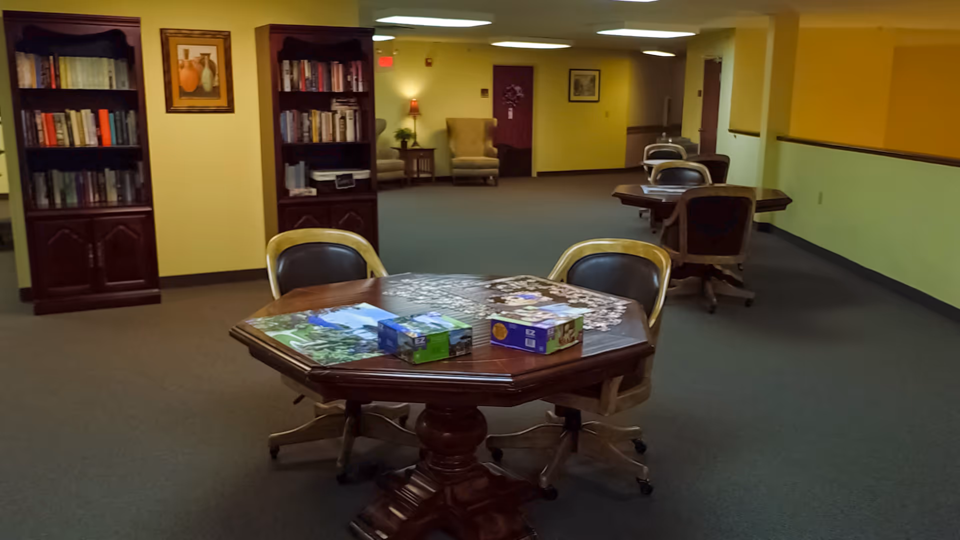 A common area in a senior living facility with a wooden octagonal table in the foreground, partially completed puzzles on the table, and four chairs around it. In the background, there are two wooden bookshelves filled with books, a framed picture on the wall, and additional tables and chairs along the hallway. The walls are painted yellow and green, and the floor is carpeted.
