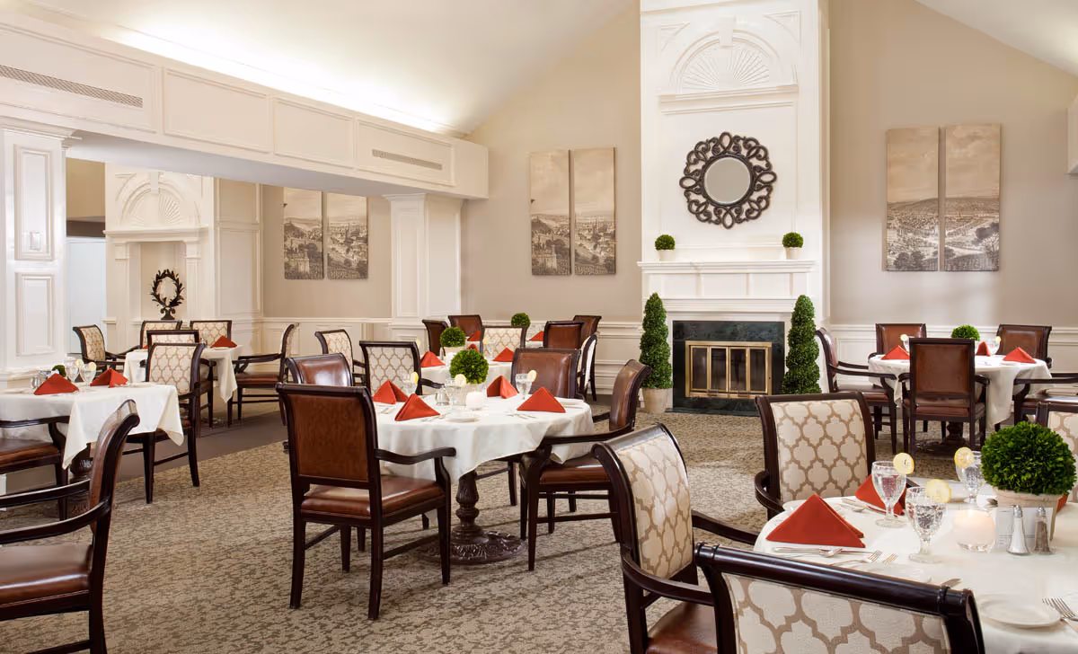 Elegant dining room with round tables set with white tablecloths and red napkins, upholstered chairs, and a fireplace in the background.