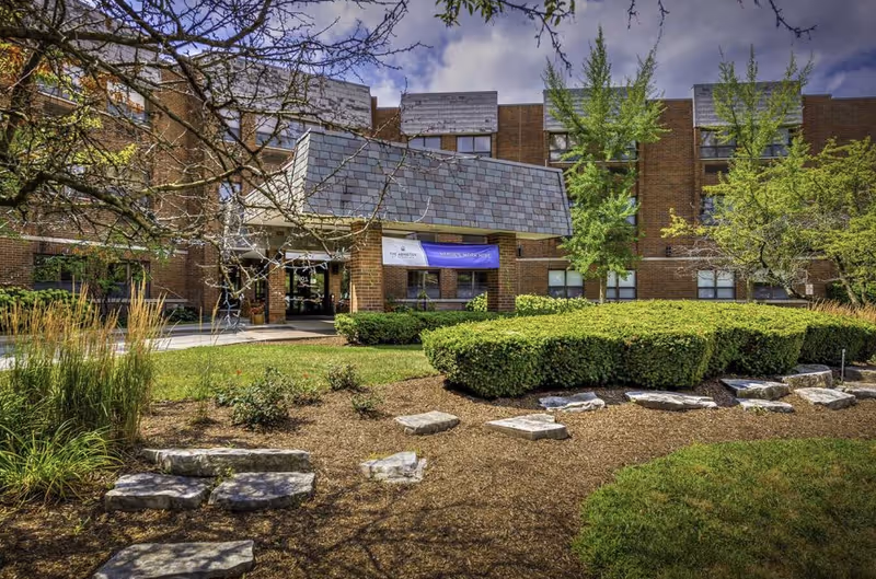 Exterior view of a brick senior living facility building with a covered entrance. The foreground features landscaped bushes, trees, and a stone pathway leading to the entrance. The sky is partly cloudy.