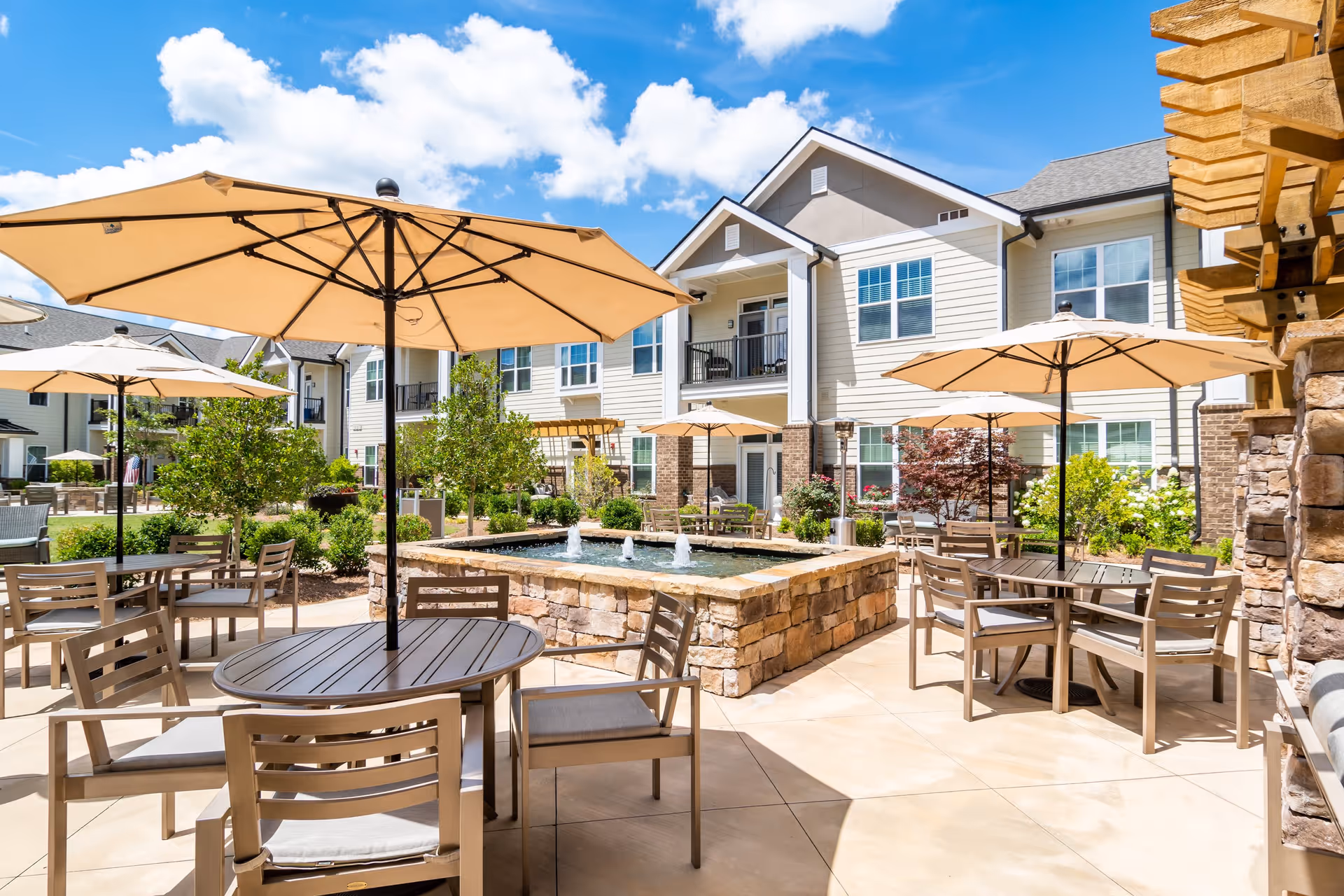 Outdoor patio area at Arbor Terrace Hamilton Mill with multiple tables and chairs under large beige umbrellas, a stone water fountain in the center, surrounded by greenery and a two-story residential building under a bright blue sky with scattered clouds.