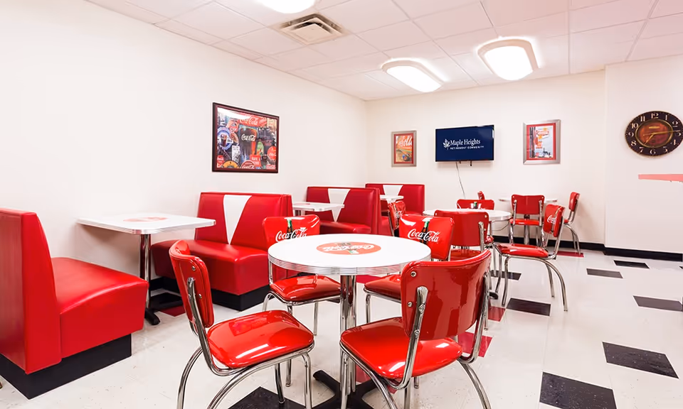A retro-style dining room with red vinyl booths and chairs featuring Coca-Cola branding. The room has white walls, a black and white checkered floor, and framed Coca-Cola posters on the walls. A flat screen TV on the wall displays the Maple Heights Senior Living logo.