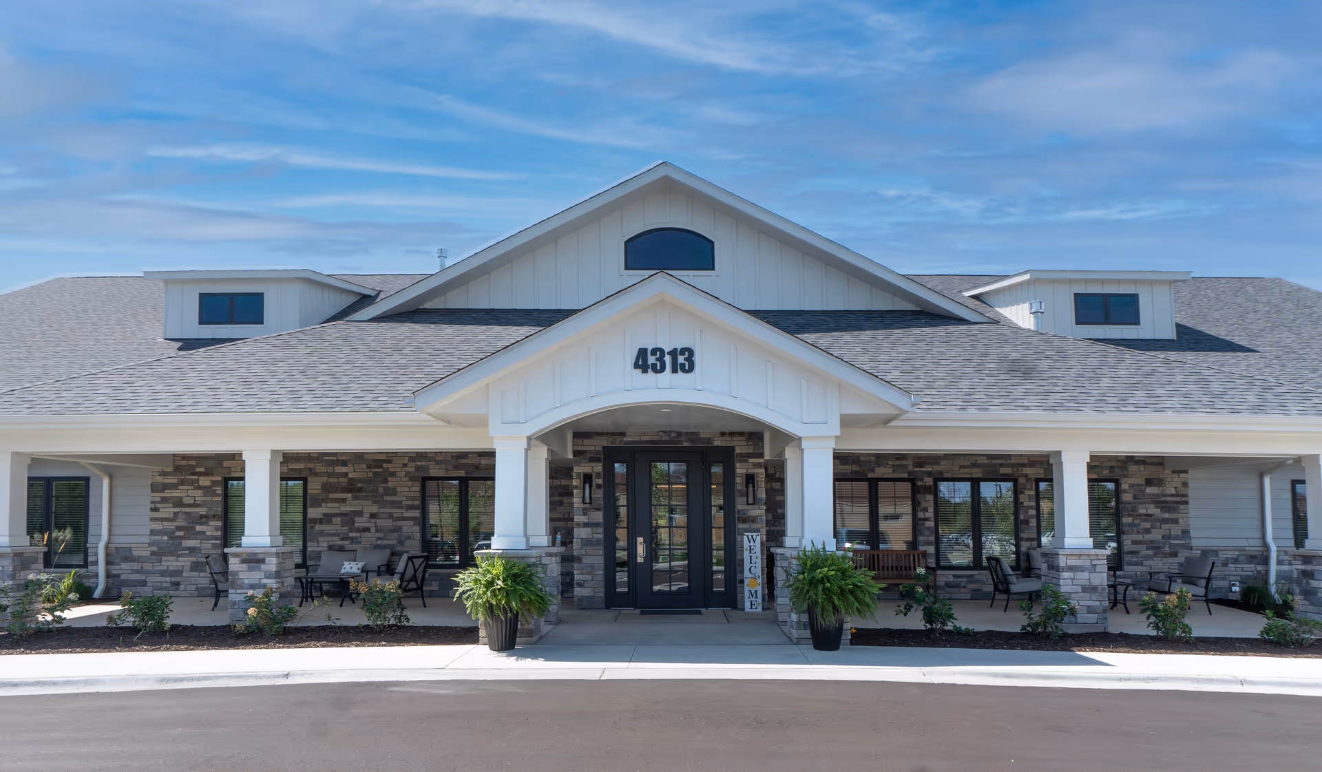 Front exterior view of a single-story building with stone and white siding, a covered entrance with the number 4313 above the door, two large potted plants flanking the entrance, and outdoor seating areas on both sides under the porch.