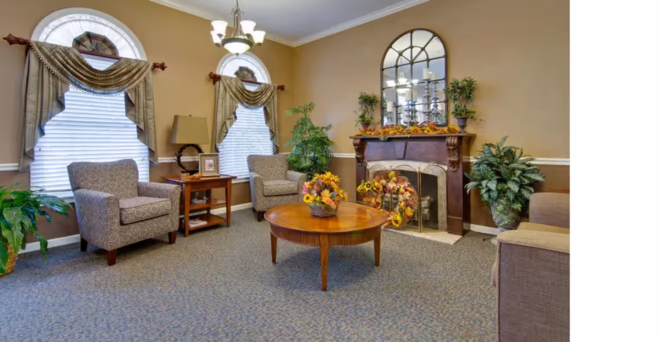 Cozy common room with armchairs around a round coffee table, a decorated fireplace with a mirror, and potted plants.