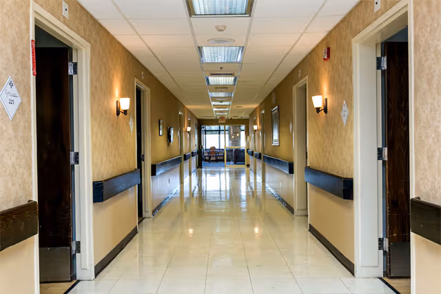 Long, well-lit nursing home corridor with open resident rooms on both sides and a seating area visible at the far end.