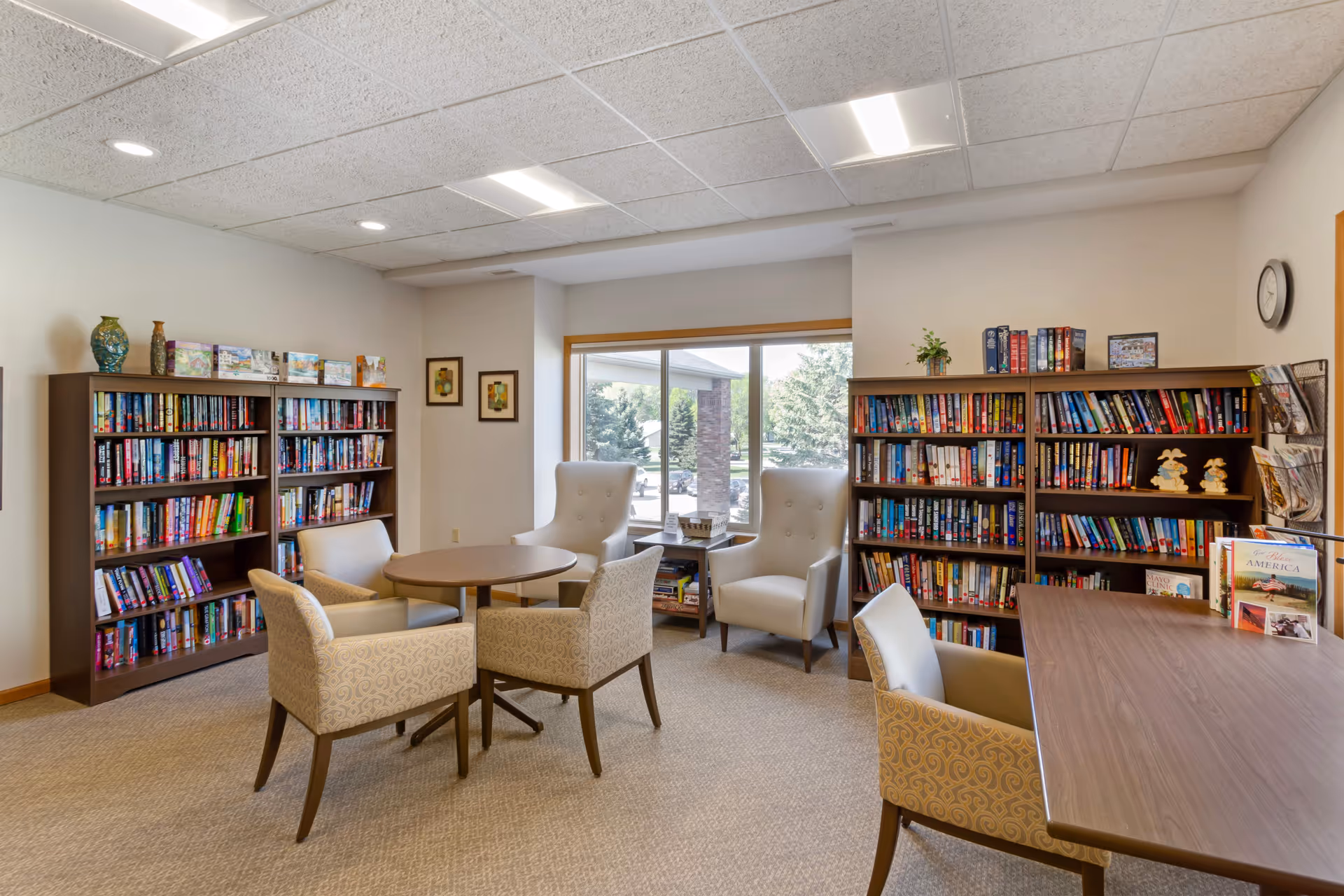 A cozy senior living facility library room with bookshelves filled with books along the walls, a round table surrounded by four upholstered chairs, two high-back armchairs near a large window, and a rectangular table with a chair. The room is well-lit with ceiling lights and has a carpeted floor.