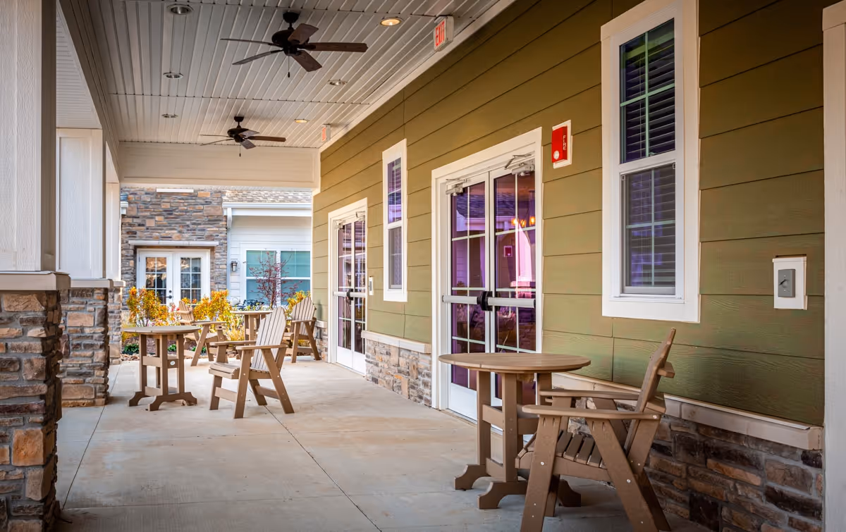Covered outdoor patio area with wooden tables and chairs, ceiling fans, and green siding walls with white trim. Stone accents are visible on the lower part of the walls and columns. Glass doors and windows lead inside the building.