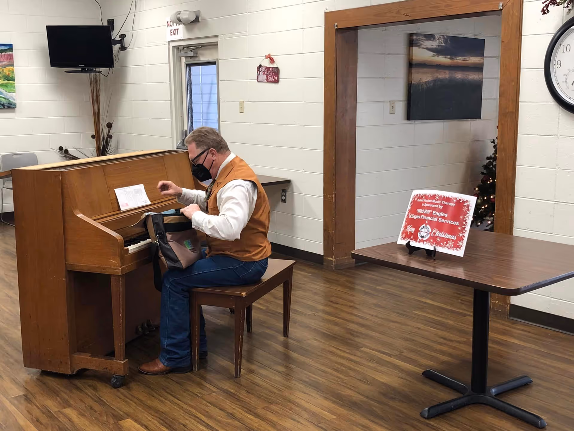 A man wearing a mask and a brown vest is sitting on a wooden bench playing an upright piano in a room with wooden flooring and white brick walls. There is a table with a Christmas-themed sign, a clock on the wall, and a small decorated Christmas tree in the background.