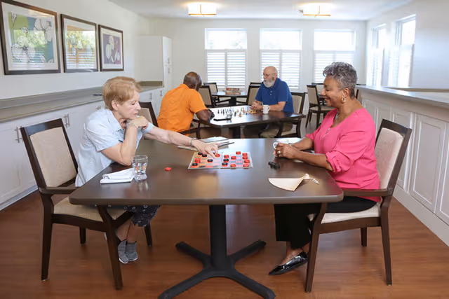 Two elderly women sitting at a table playing a board game in a well-lit room with large windows. In the background, two elderly men are seated at another table playing chess. The room has wooden floors, white walls with wainscoting, and framed artwork on the walls.