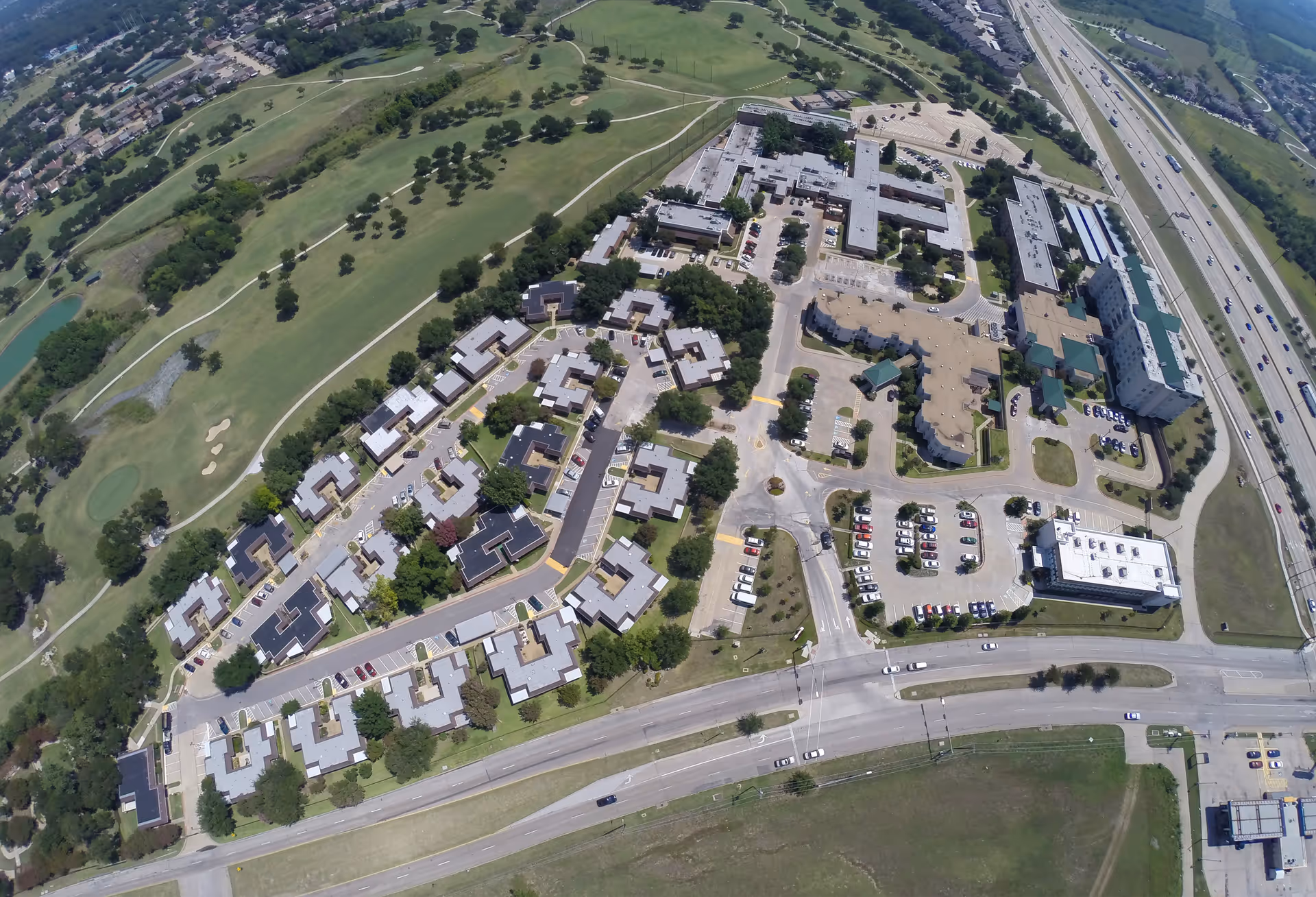 Aerial view of a senior living and rehabilitation campus with multiple buildings, parking areas, an adjacent golf course, and a nearby highway.