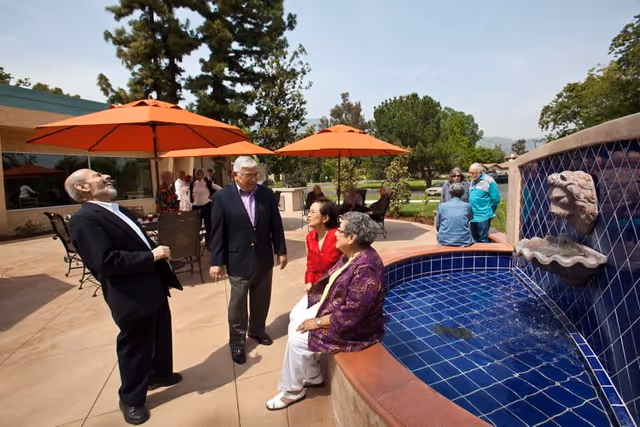 A group of elderly people socializing outdoors near a blue-tiled water fountain with a lion head spout. Two orange umbrellas provide shade over patio tables and chairs. Trees and greenery are visible in the background under a clear sky.